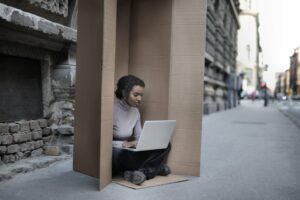Young female introvert in casual clothes typing on netbook while sitting with crossed legs inside cardboard box located on as