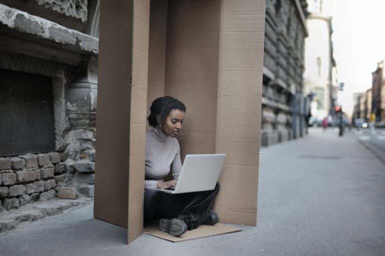 Young female introvert in casual clothes typing on netbook while sitting with crossed legs inside cardboard box located on as