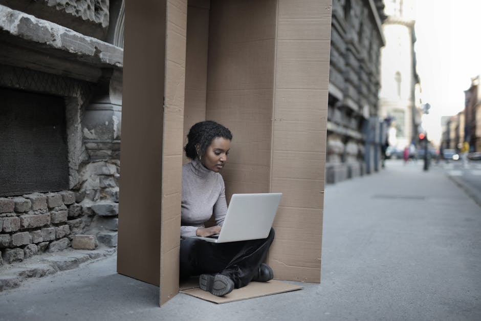Young female introvert in casual clothes typing on netbook while sitting with crossed legs inside cardboard box located on as