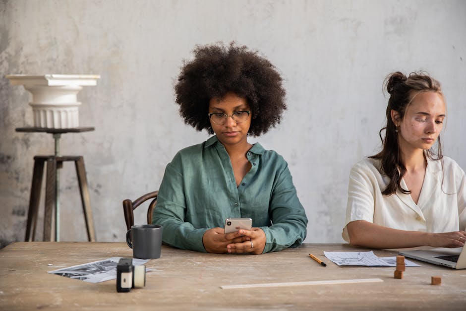 Two women in a modern office using smartphone and laptop at a shared desk.