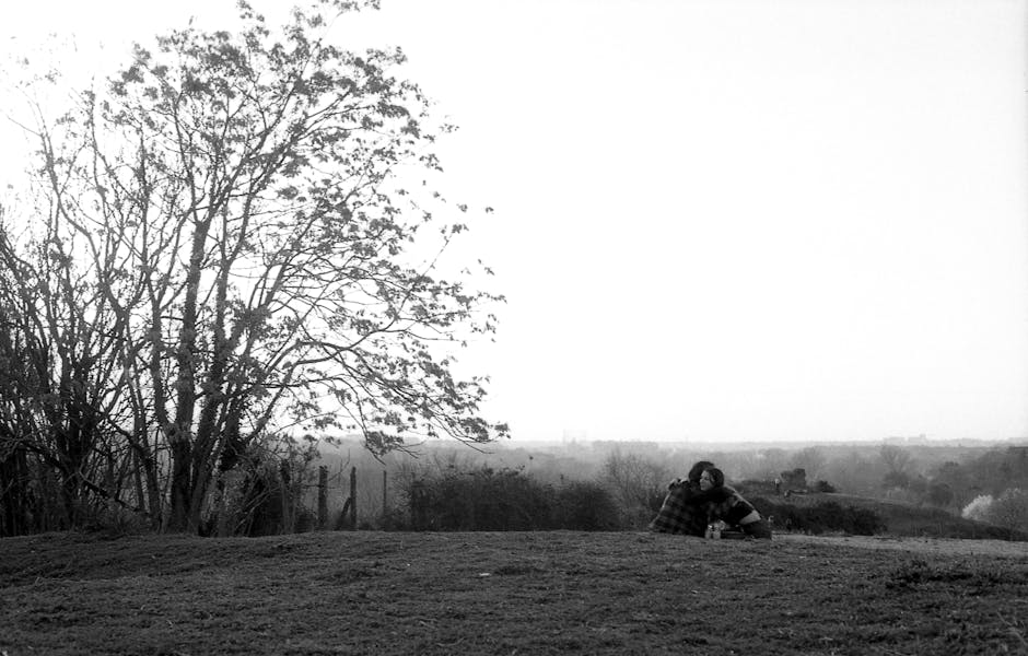 Black and white of anonymous woman embracing boyfriend while resting on lawn against trees in daytime