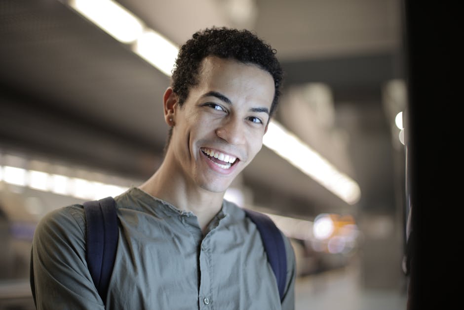 Low angle of joyful charismatic African American male in gray shirt with backpack smiling at camera while standing against bl