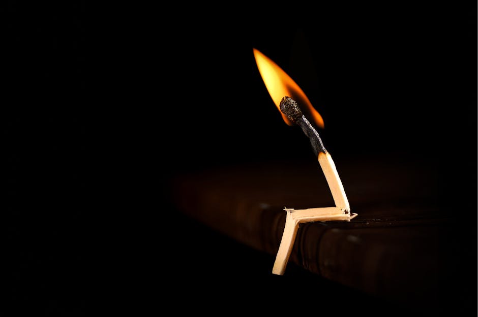A single matchstick burning on a dark surface, casting a warm glow.
