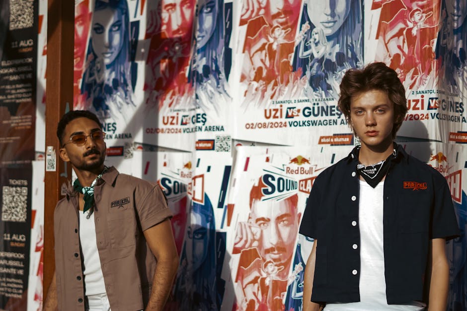 Two men pose in fashion outfits against vibrant urban event posters in daylight.