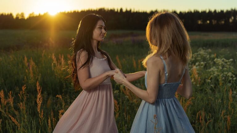 Two women in dresses holding hands in a sunlit field at sunset.