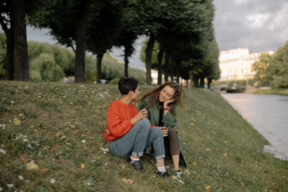 Two women enjoying coffee and conversation while sitting by a river in a picturesque autumn park.