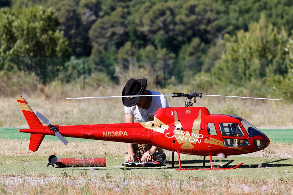 A man adjusts a model helicopter on a grass field with a forest background.