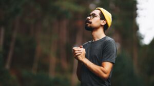 A man looks up thoughtfully in a forest, wearing a yellow beanie and glasses.