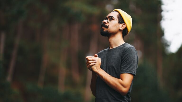 A man looks up thoughtfully in a forest, wearing a yellow beanie and glasses.