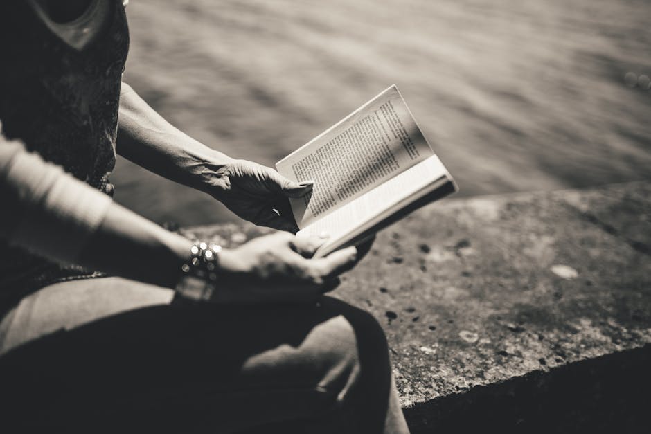 A woman enjoys reading a book outdoors by the water in a monochrome setting.