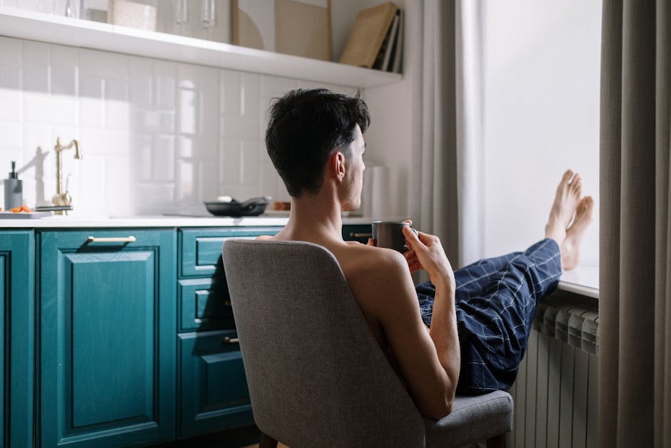 Man sitting on a chair with coffee, legs on windowsill, in a kitchen.