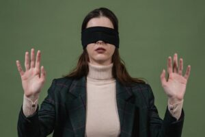 Blindfolded woman standing with hands raised against a green background.