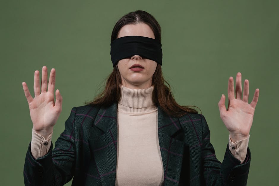 Blindfolded woman standing with hands raised against a green background.