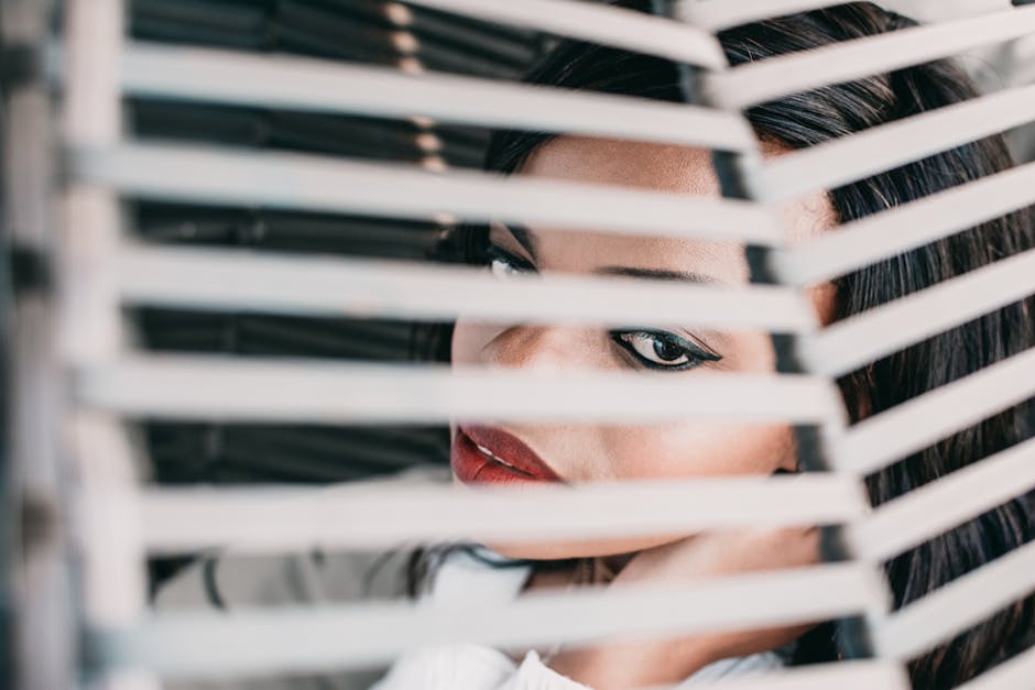 Close-up of a stylish woman peering through window blinds indoors.