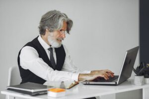 Elegant senior man in office attire typing on a laptop at a desk.