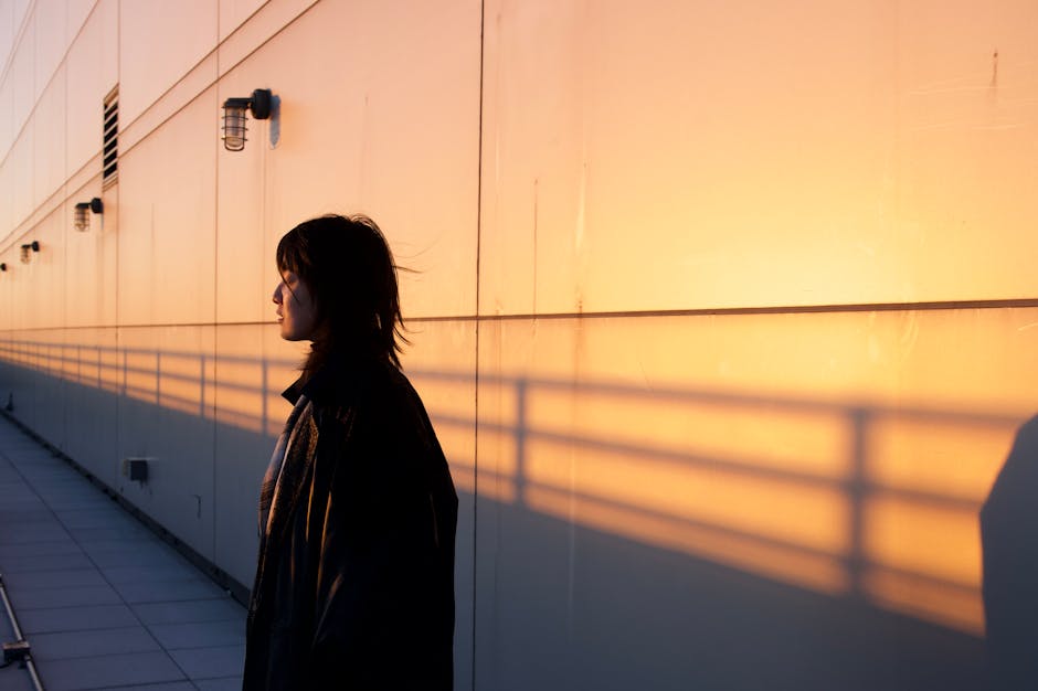 A woman stands in silhouette on a city rooftop as the sun sets, casting warm shadows on a building facade.