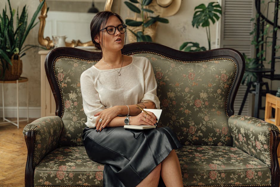 A woman sitting on a vintage floral couch indoors, holding a notebook and wearing eyeglasses.