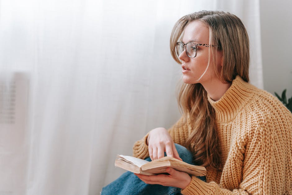 Young female in casual clothes and glasses sitting and holding book while looking away near window with white curtain