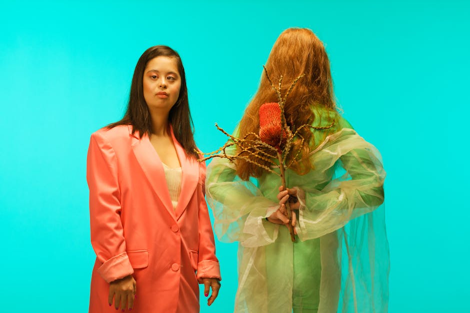 Two women posing against teal background, one in a pink blazer, the other holding a dried flower.