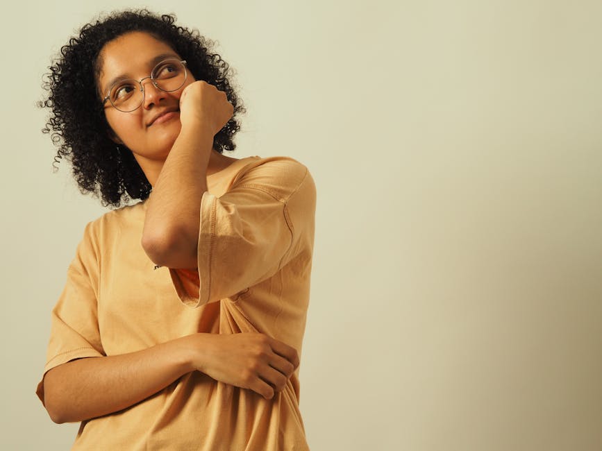 Thoughtful person with curly hair and glasses in a casual pose against a neutral background.
