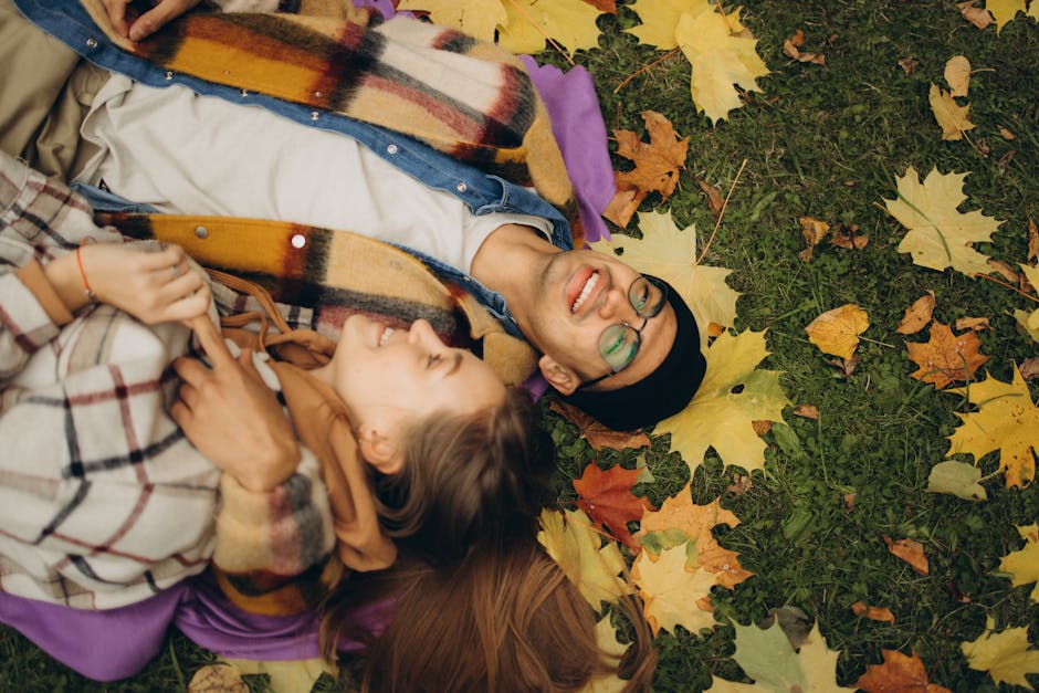 Smiling couple lying on colorful autumn leaves, enjoying a warm, joyful moment outdoors.