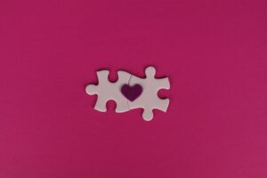 Two white puzzle pieces with a heart symbol on a pink background, symbolizing love and connection.
