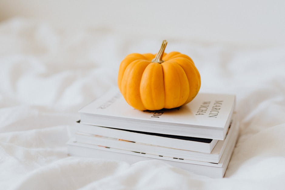 A bright orange pumpkin rests on a stack of books, set against a soft white background.
