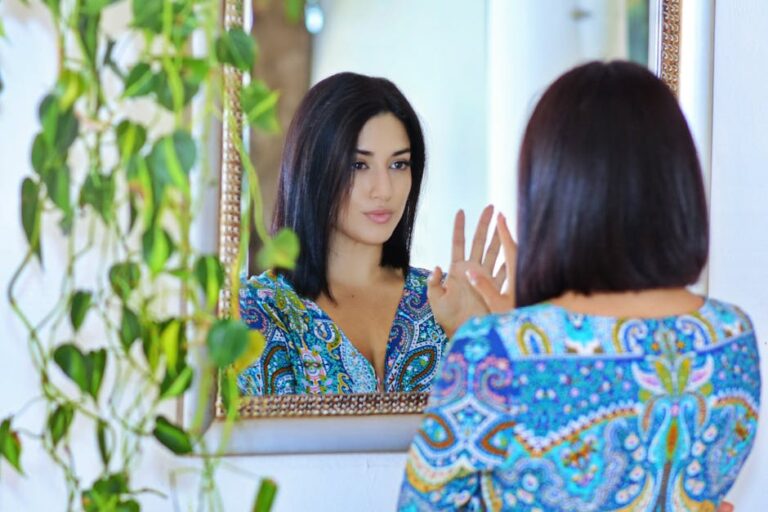 A woman in a vibrant patterned shirt gazes at her reflection in a mirror surrounded by green plants.