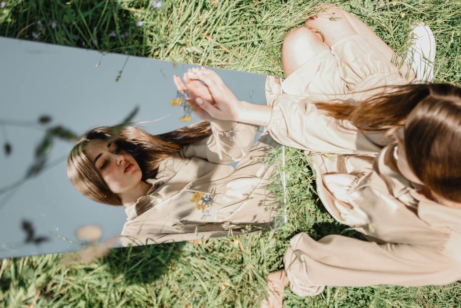 A woman in a beige outfit sits in grass, reflected in a mirror outdoors.