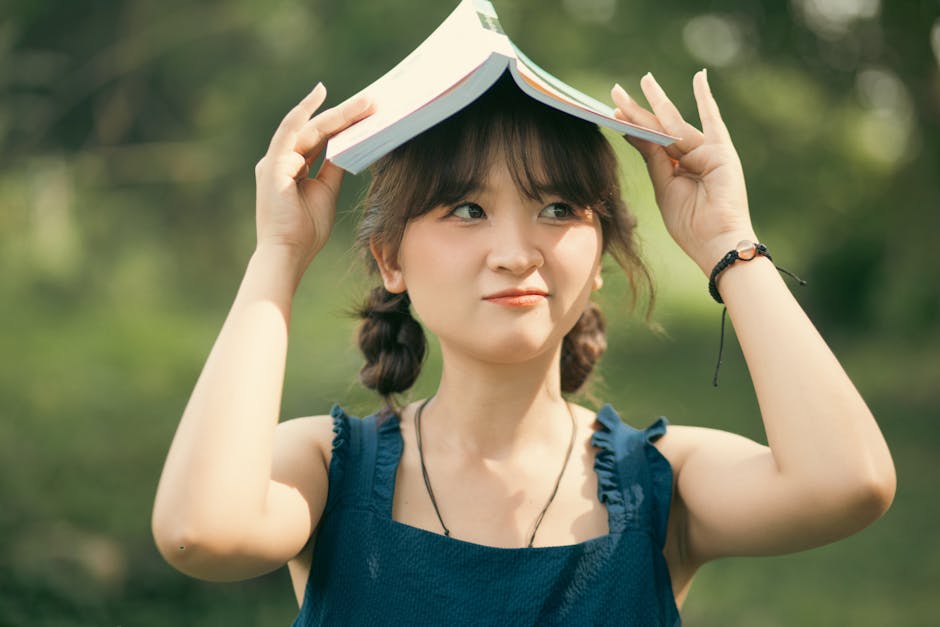 Playful young woman with a book on her head smiling outdoors.