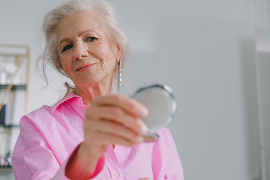 Elegant elderly woman in pink shirt holding a compact mirror indoors, reflecting joy and confidence.