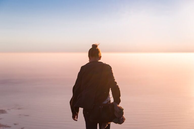 A woman in a black jacket stands at the seashore in Cape Town, admiring a tranquil sunset over the ocean.