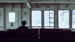 Person sitting alone on a ferry, facing the sea through large windows, reflecting solitude.