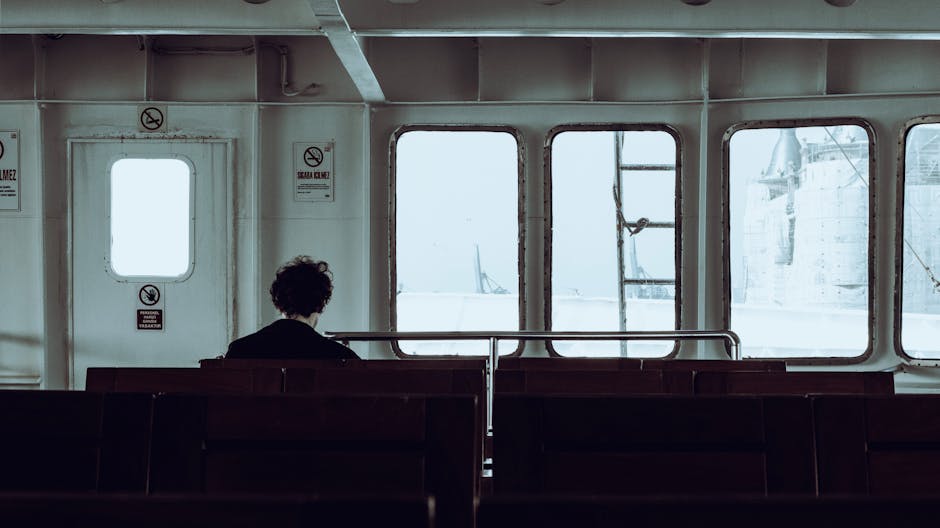 Person sitting alone on a ferry, facing the sea through large windows, reflecting solitude.