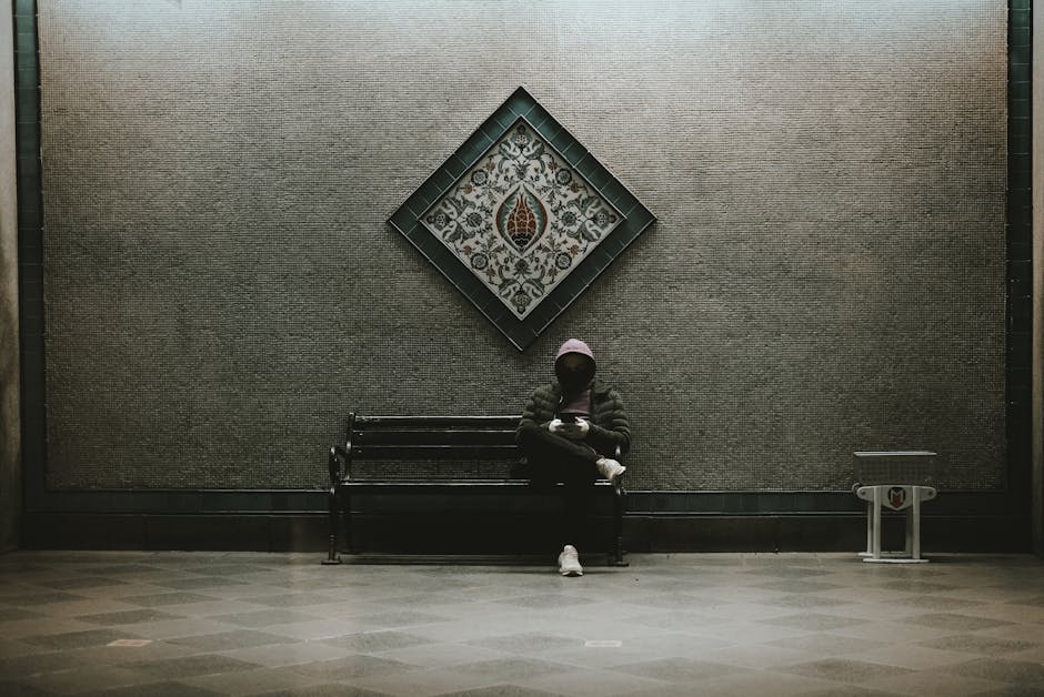 A person in a hoodie sits on a metal bench beneath decorative wall art, capturing a moody, urban scene.