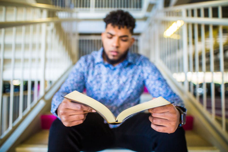 Focused young man reading a book on a staircase indoors, creating a study or learning vibe.