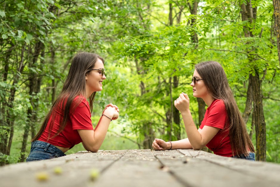 Two young women smiling and chatting in a vibrant green forest while sitting at a wooden table.