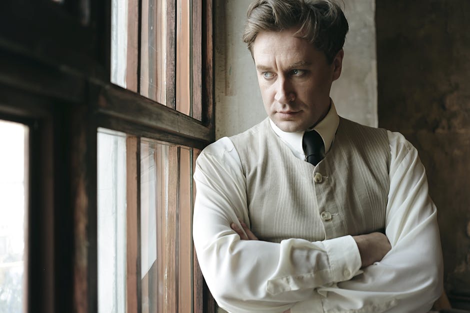 A thoughtfully dressed man in vintage attire gazing outside through a window, creating a nostalgic atmosphere.