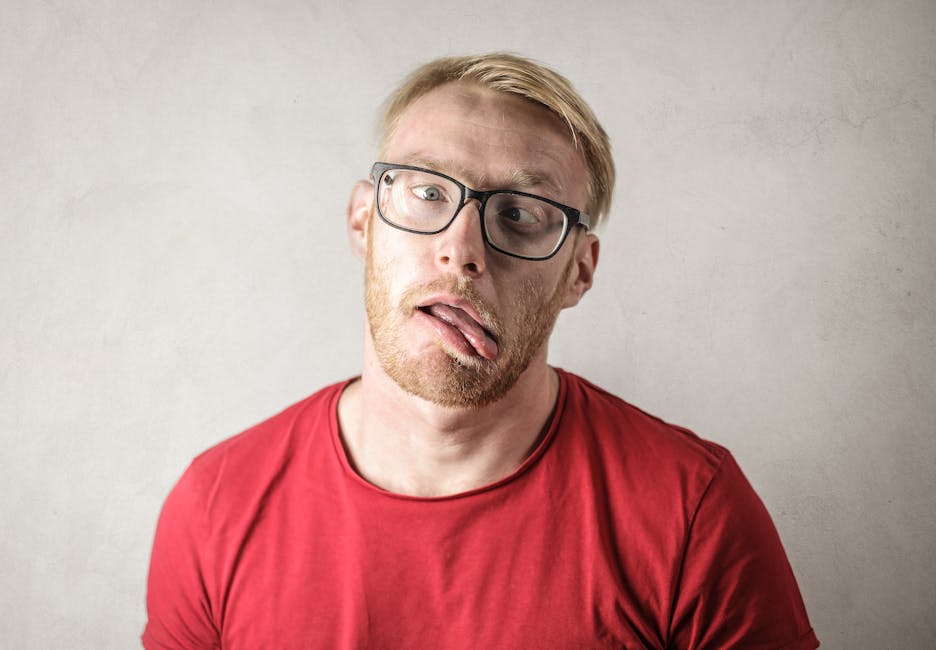 Man in a red shirt making a funny face with tongue out, wearing glasses.