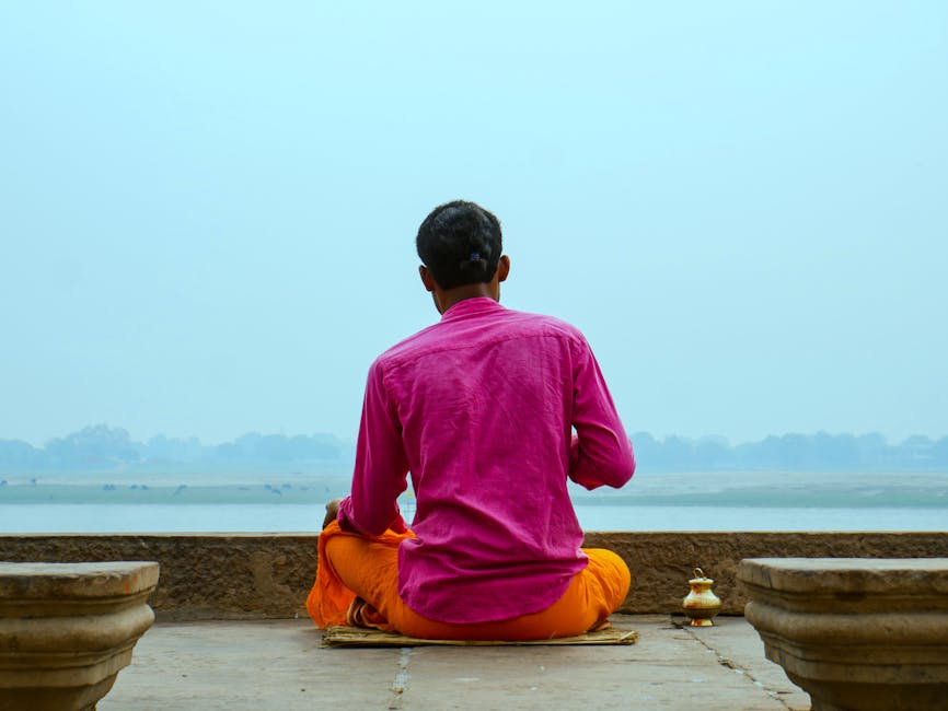A man in colorful attire meditates by the river, enjoying peaceful solitude.