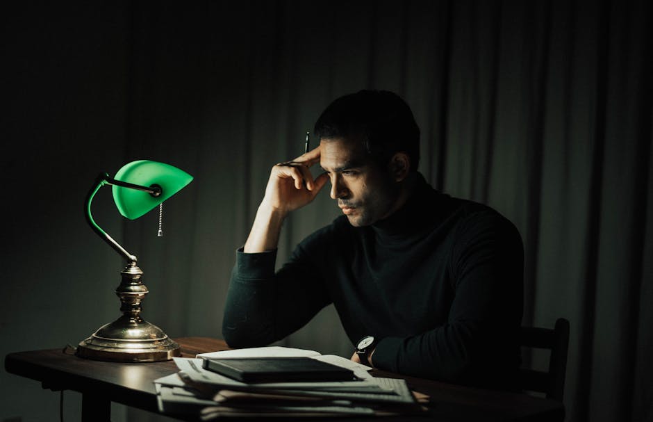 Side view of pensive male with pen doing paperwork while sitting at wooden desktop with vintage table lamp during work in obs