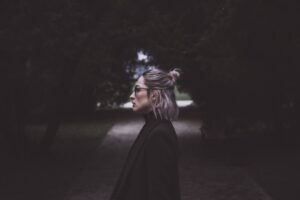 Profile of a fashionable woman with glasses walking down a dimly lit pathway, surrounded by trees.