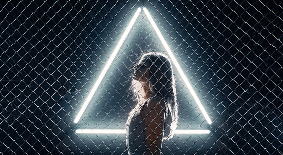 Side profile of a woman behind a chain-link fence with a glowing triangle, creating a striking silhouette.
