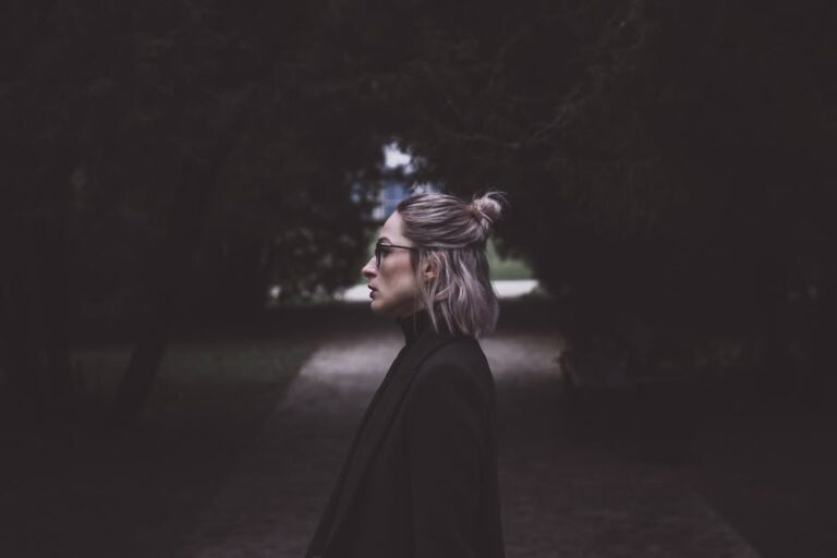 Profile of a fashionable woman with glasses walking down a dimly lit pathway, surrounded by trees.