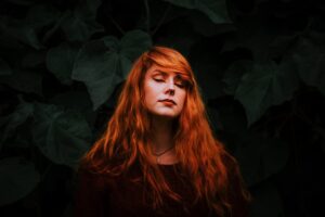 A calm redhead woman with eyes closed stands against lush dark green leaves, embodying serenity in nature.