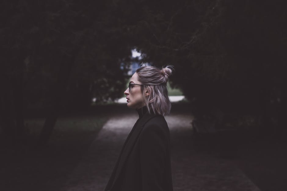 Profile of a fashionable woman with glasses walking down a dimly lit pathway, surrounded by trees.