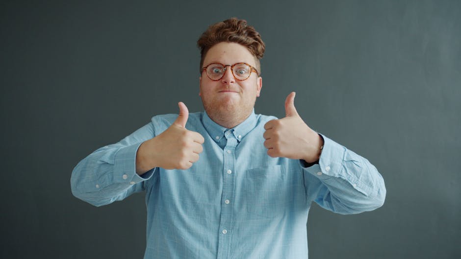 Smiling adult in glasses and blue shirt showing thumbs up indoors.
