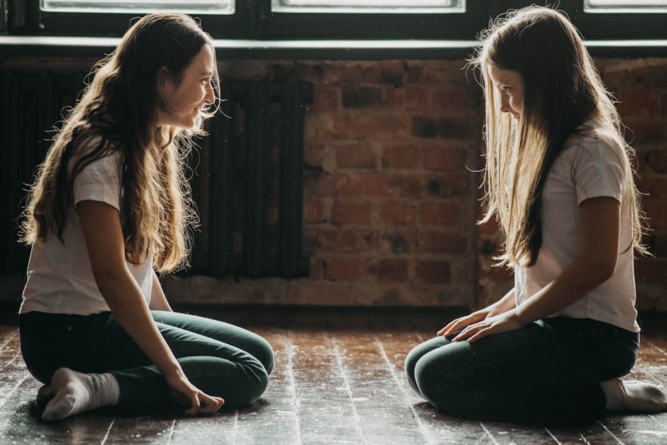 Two teenage girls are kneeling on a wooden floor, smiling and talking face to face indoors.