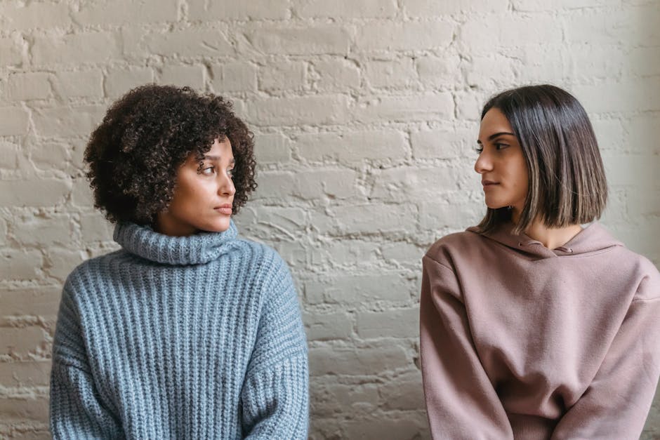 Emotionless young multiethnic women in casual outfit looking at each other near white brick wall in bright house