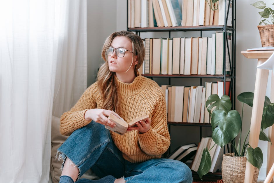 Pensive young female in casual outfit and eyeglasses sitting on floor near bookshelf and plants holding book while looking at